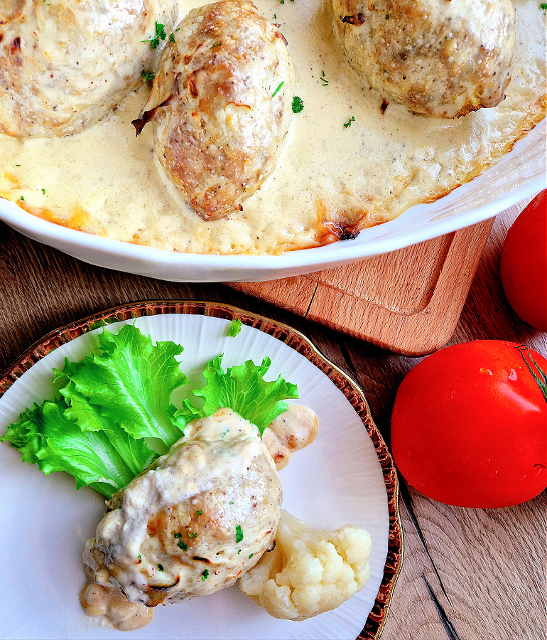 Keto gołąbki bez zawijania podane na biało-złotym talerzyku. Obok na desce naczynie żarodporne z gołąbkami w sosie. Obok pomidor.