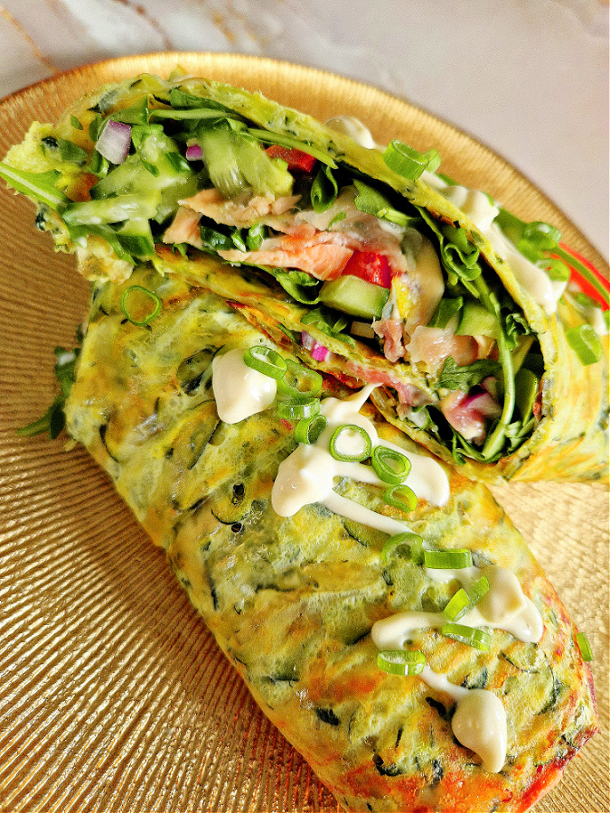 Wrap z cukinii nadziewany rukolą, łososiem, sosem czosnkowym, czerwoną cebulą i pomidorami na złotym talerzu