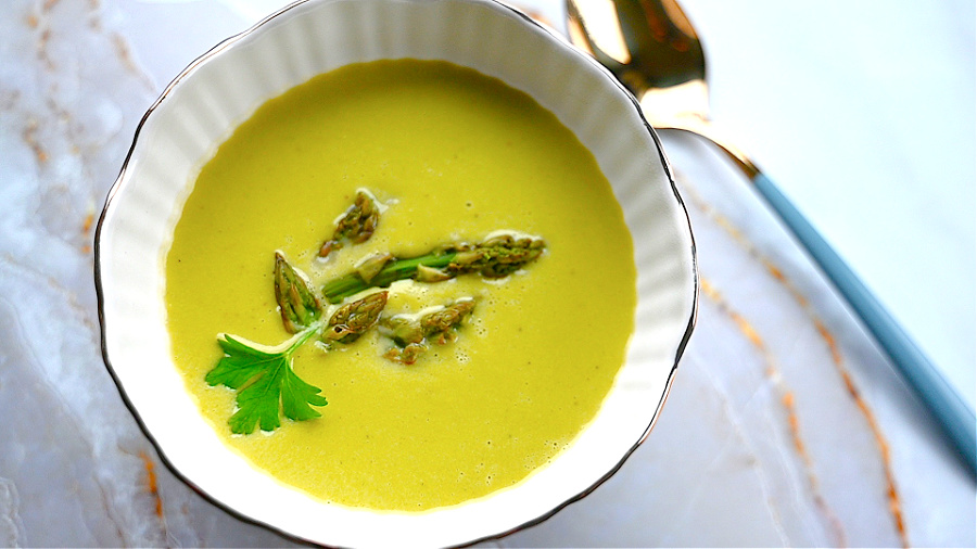 Biała miska ze złotą obwódką, a w niej aksamitna zupa z zielonych szparagów z kawałkami szparagów i zieloną pietruszką. Tło biało-złote.