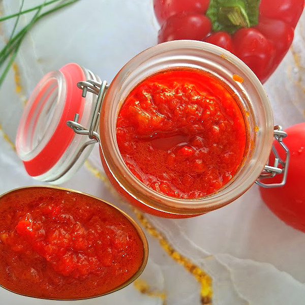 Zdjęcie główne kwadratowe. Szklany słoik zamykany na klamrę wypełniony pastą ajvar, obok złota łyżka z porcją pasty, w tle papryka, pomidory i świeża zielenina.