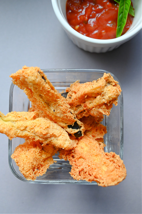 Przezroczysta miseczka z usmażonymi w tempurze keto paskami bakłażana, obok biała miseczka z sosem pomidorowym z bazylią, całość na szarym blacie.