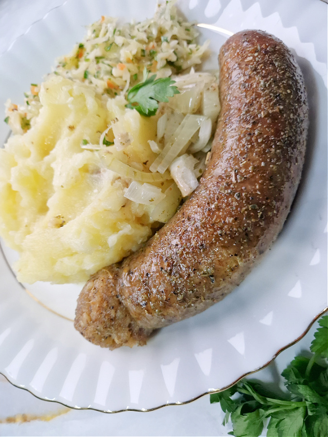 Biała kaszanka z piekarnika z cebulką podana na białym talerzu z ziemniakami puree i surówką z kiszonej kapusty.