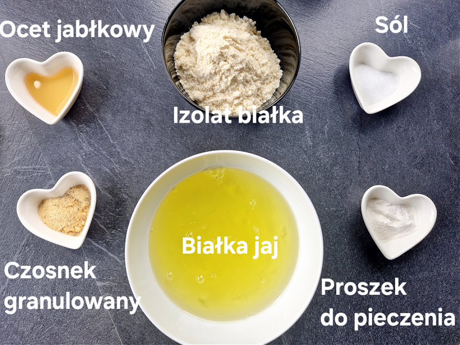Składniki do przygotowania keto bułki tartej ułożone w miseczkach na ciemnym blacie. Izolat białka, białka ja, proszek do pieczenia, czosnek granulowany, ocet jabłkowy, sól