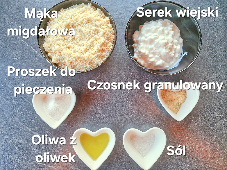 Składniki na keto bułeczki: serek wiejski, mąka migdałowa, jajko, proszek do pieczenia, olej, czosnek granulowany, w miseczkach na ciemnym blacie.