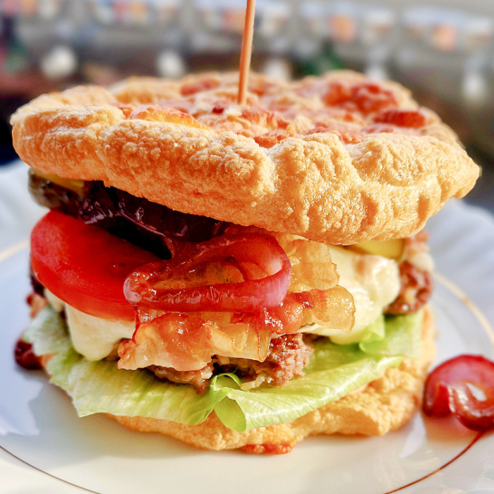 Zbliżenie na gotowego keto burgera z wołowiną, podanego na białym talerzu. W środku widać sałatę, soczystą wołowinę z sosem, plasterki pomidora, ogórka i czerwoną cebulę, przykryte bułką keto. W tle stoją słoiczki z przyprawami.