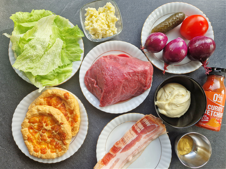 Składniki na keto burgera rozłożone na talerzach i w miseczkach na antracytowym blacie. Widać mięso wołowe, domowe bułeczki keto, zieloną sałatę, ser, majonez, musztardę, sos bez cukru, czerwoną cebulę, ogórki, pomidora i plastry boczku.