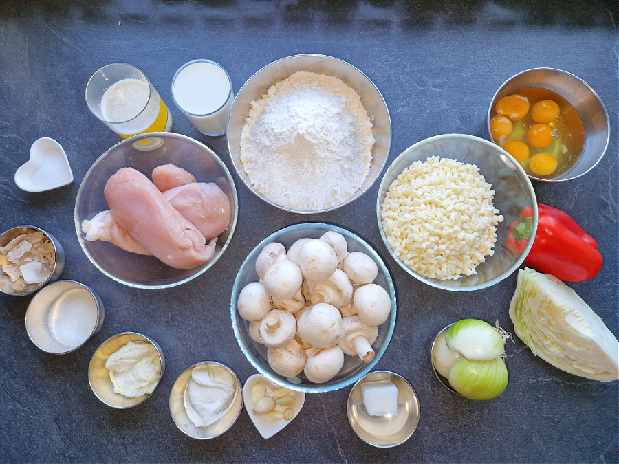 Na ciemnym blacie ustawione są wszystkie składniki potrzebne do calzone z kurczakiem. W miseczkach i na talerzykach leżą: surowe filety z kurczaka, pieczarki, mąka, starta mozzarella, jajka, papryka, ćwiartka kapusty białej, cebula, kostka smalcu, ząbki czosnku, jogurt, majonez, sól, świeże drożdże, masło i szklanka mleka.