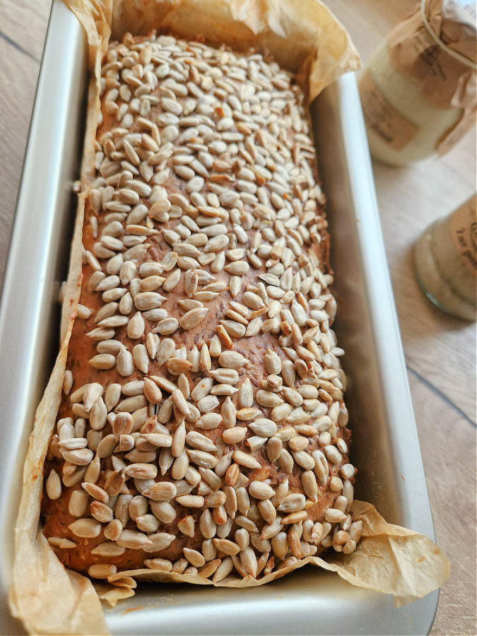 Bochenek chleba twarogowego low carb z chrupiącą skórką prosto po wyjęciu z piekarnika. Pieczony w brytfance wyłożonej papierem do pieczenia. Blat drewniany, obok słoiczki.