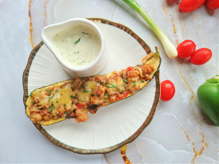 Biało-złoty talerz na jasnozłotym tle. Na nim zapieczona połówka cukinii nadziewana mięsem mielonym i serem. Obok sosjerka z sosem czosnkowym, kilka pomidorków koktajlowych i cebulka dymka.