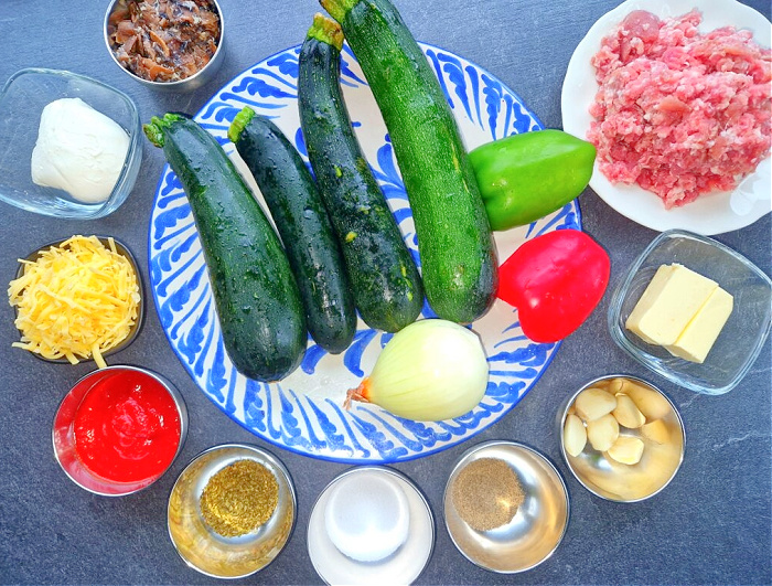 Ciemny antracytowy blat, na którym ułożone są wszystkie składniki do faszerowanej cukinii: świeże cukinie, mięso mielone, ser mozzarella, ser żółty gouda, czerwona i zielona papryka, pieczarki, cebula, masło, passata pomidorowa, czosnek oraz przyprawy t.j. sól, pieprz i mieszanka włoska do pizzy.