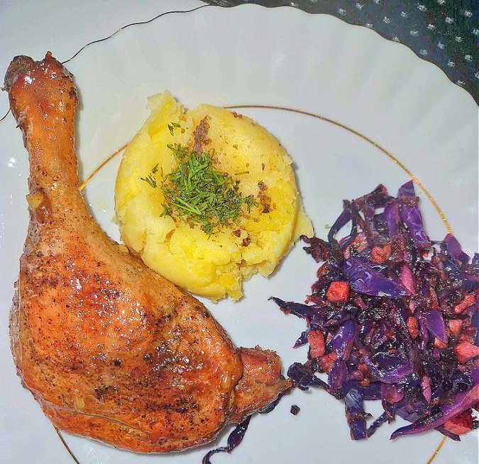 Biały talerz ze złotą obwódką, na którym podane są ziemniaki, udko z kaczki oraz porcja czerwonej kapusty duszonej z boczkiem i cebulką.