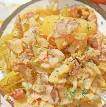 Zbliżenie na talerz z duszonkami przygotowanymi w Instant Pot. Ziemniaki, kapusta, mięso i kiełbasa, udekorowane świeżą natką pietruszki, na jasnym tle.