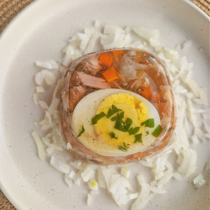 Kwadratowe zbliżenie na porcję galaretki z nóżek i golonki. W środku jajko, kawałki mięsa i warzyw. Obok na talerzyku leży posiekana cebula. Tło brązowe.