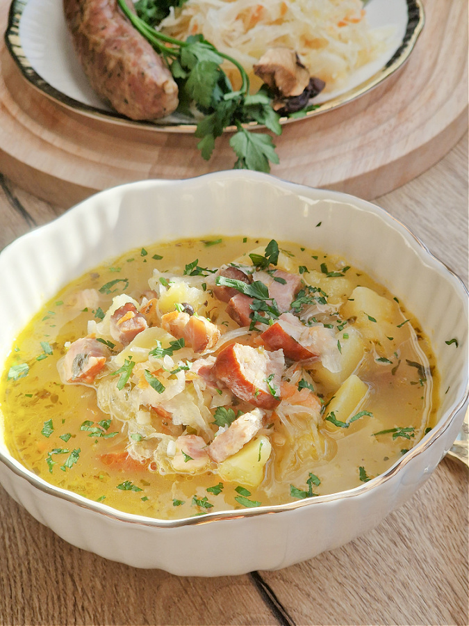 Zdjęcie kapuśniaku z kiszonej kapusty który jest w biało złotej misce. W tle talerzy z kiełbasą, natką pietruszki i kapustą kiszoną. Blat drewniany.