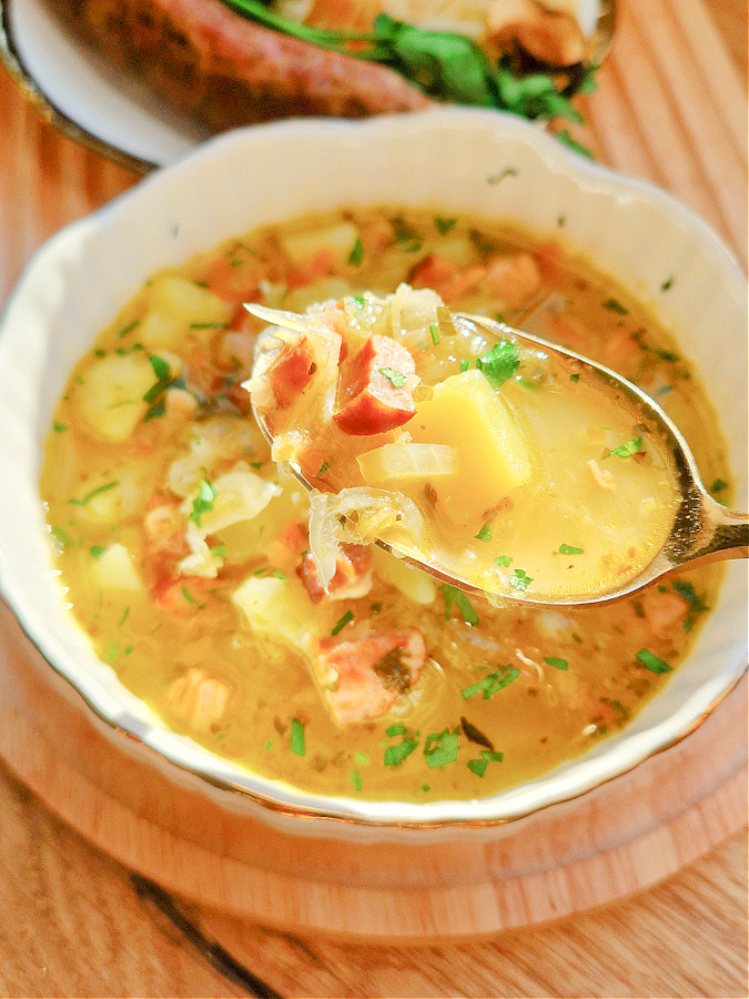 Kapuśniak z kiszonej kapusty z ziemniakami i kiełbasą w białej miseczce, nałożony na łyżkę. W tle drewniany blat.