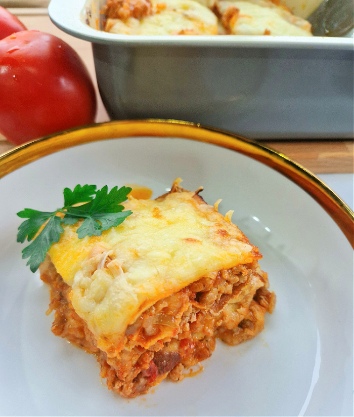 Porcja keto lazanii zapiekanej z serem w białej miseczce ze złotą obwódką, obok brytfanka z lazanią i pomidor.