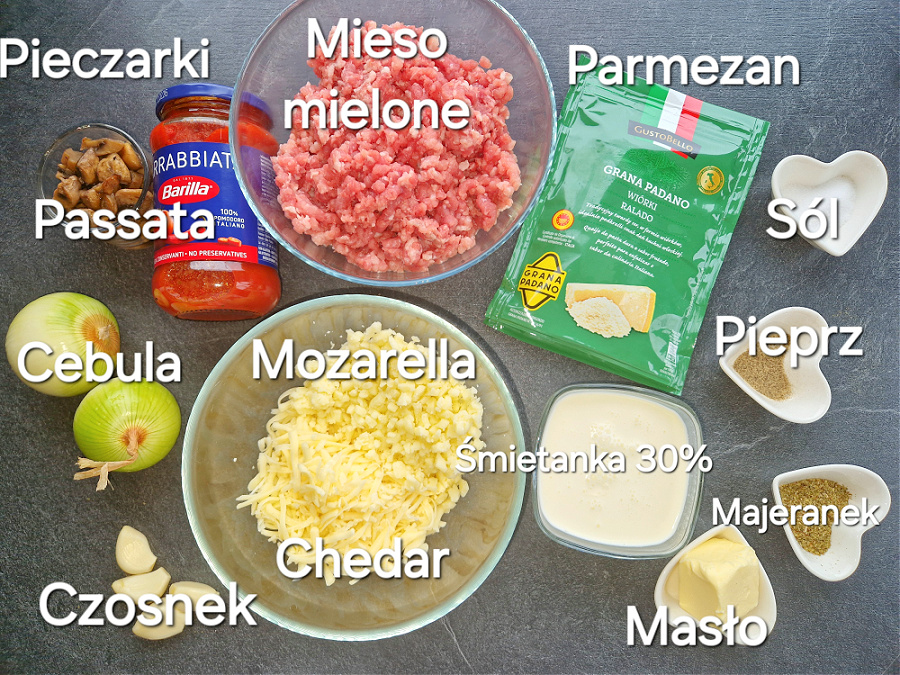 Składniki na keto lazanię: pieczarki, passata, cebula, czosnek, mozzarella, cheddar, mięso mielone, parmezan, śmietanka, masło i przyprawy na ciemnym blacie.