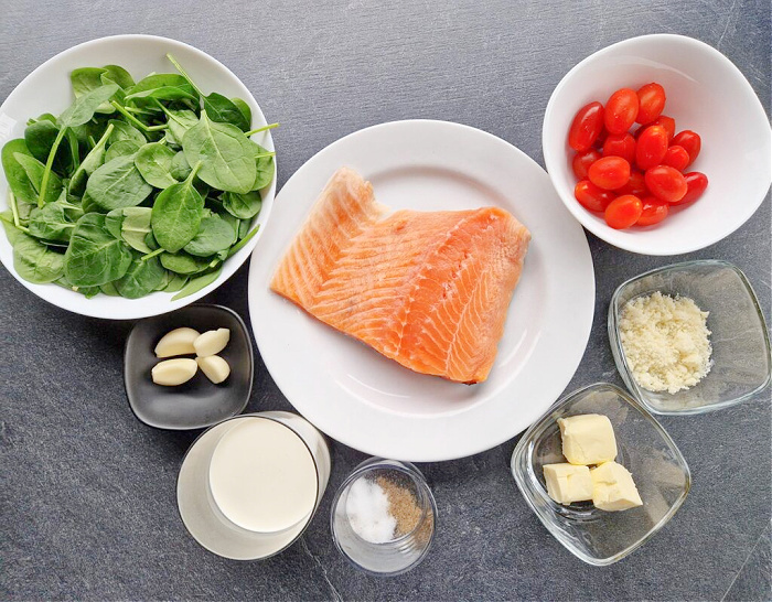 Biały talerz z porcją surowego fileta z łososia, obok w miseczkach przygotowane składniki: świeży szpinak, czosnek, śmietanka, przyprawy, masło, tarty ser oraz czerwone pomidorki koktajlowe. Tło to ciemny blat.