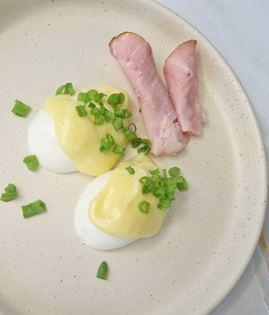 Kremowy talerz z dwiema połówkami jajka ugotowanego na twardo, polanymi domowym majonezem z żółtek i posypanymi świeżym szczypiorem. Obok znajdują się zwinięte plastry szynki. Całość podana na jasnym tle.