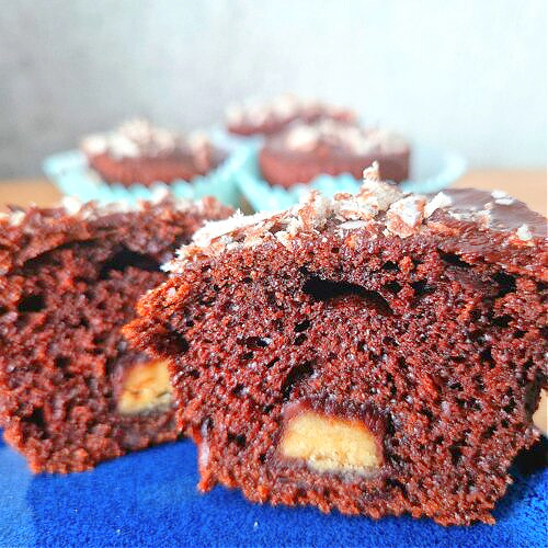 Kwadratowe zdjęcie główne. Ujęcie z boku na przekrojoną muffinkę czekoladową. W środku widoczna czekoladka Merci. Wierzch polany czekoladą i posypany kruszonym Prince Polo. W tle kilka muffinek na niebieskim talerzu.