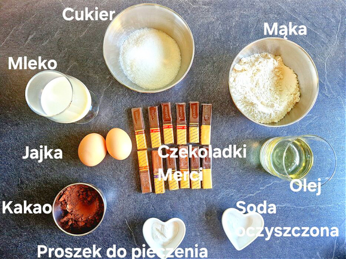 Na ciemnym antracytowym blacie ułożone składniki do muffinek: w szklankach i miseczkach kakao, mąka, cukier, proszek do pieczenia, soda, olej i mleko. Obok jajka i czekoladki Merci.