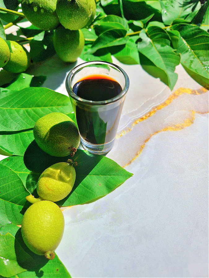 Kieliszek wypełniony ciemną, aromatyczną nalewką z zielonych orzechów, ustawiony na jasnozłotym tle. Obok kieliszka świeże liście i zielone orzechy włoskie.