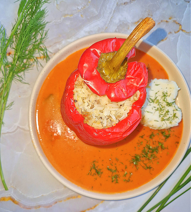 Jasny kremowy talerz z sosem pomidorowym, na którym leży papryka faszerowana mięsem i ryżem, przykryta odciętą górną częścią. Obok serce z puree ziemniaczanego posypane koperkiem.