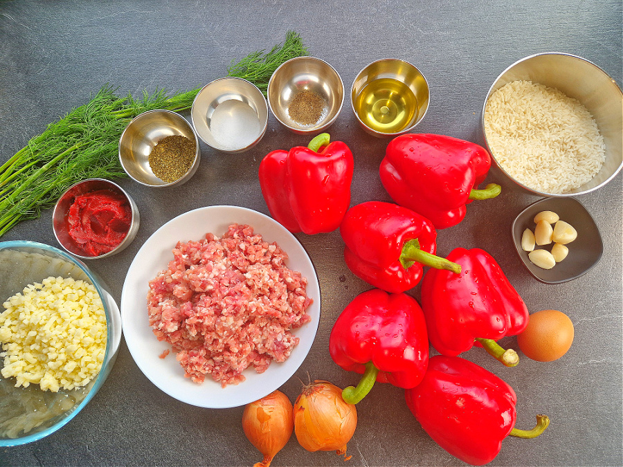 „Składniki do przygotowania faszerowanej papryki: czerwone papryki, cebula, mięso mielone, ryż, żółty ser, koncentrat pomidorowy, czosnek, przyprawy i świeża zielenina ułożone na ciemnym blacie.