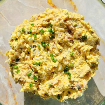 Kwadratowe zdjęcie przezroczystej miski wypełnionej pastą z makreli wędzonej i jajek. Na wierzchu posypany świeżo posiekany szczypiorek. Tło jasnozłote.