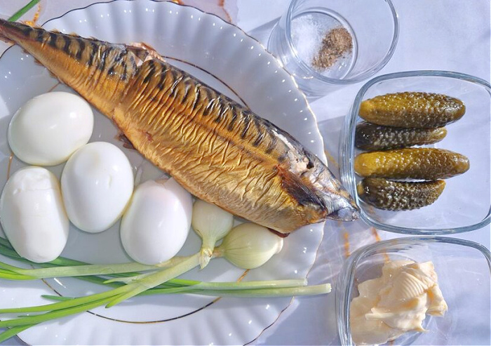 Biały talerz na jasnozłotym tle, na nim ułożona złocista makrela wędzona, obok cztery jajka ugotowane na twardo i cebulka dymka. W małych miseczkach widoczny majonez, ogórki kiszone oraz przyprawy: sól i pieprz.