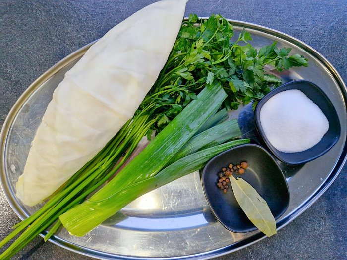 Metalowy półmisek ze składnikami przyprawiającymi rosół: zielona część pora, natka pietruszki, kawałek kapusty, a w małej miseczce ziele angielskie, liść laurowy i sól. Wszystko ustawione na ciemnym antracytowym blacie.