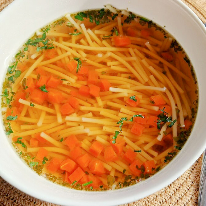 Kwadratowe zdjęcie miseczki z pieczonym rosołem. Złocista zupa z drobną marchewką w kostkę, makaronem i natką pietruszki, podana na tle ciemnego blatu.