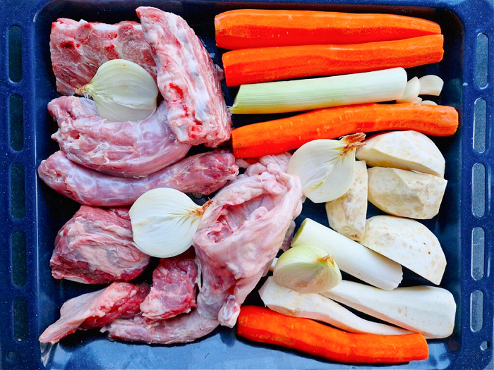 Surowe składniki ułożone na blasze do pieczenia: pokrojona marchewka, por, cebula, pietruszka, seler oraz różne kawałki mięsa. Wszystko przygotowane przed pieczeniem.