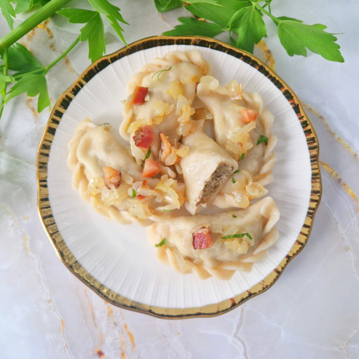 Jasny biały talerz ze złotą obwódką, na którym poukładane są pierogi z mięsem z rosołu. Całość posypana podsmażonym boczkiem i cebulką. Obok na talerzu leży świeża zielona gałązka lubczyku. Tło jasnozłote.