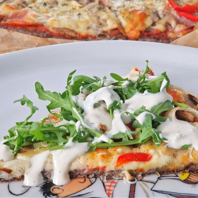 Kwadratowe ujęcie główne keto pizzy. Na pierwszym planie kolorowy talerz z kawałkiem pizzy polanej białym sosem i rukolą, w tle cała pizza na desce.