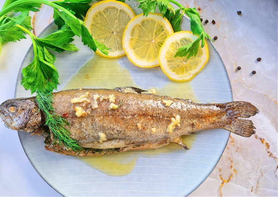 Pieczony pstrąg na błękitnym talerzu, udekorowany plastrami cytryny i zieleniną, ułożony na jasnym tle