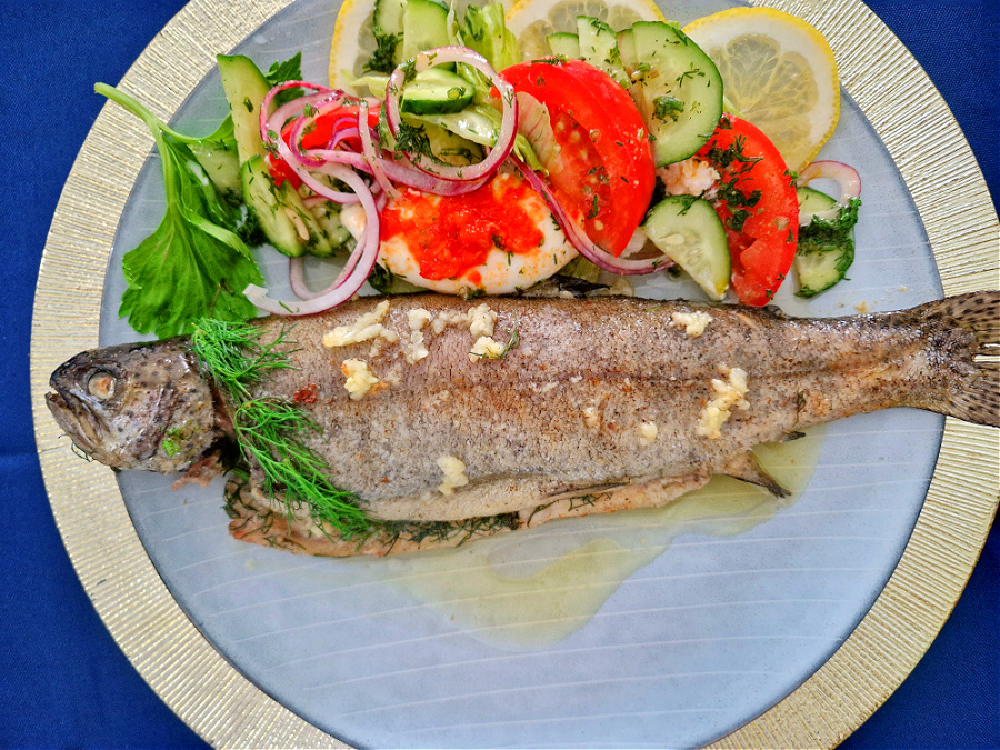 Pieczony pstrąg podany z kolorową sałatką z czerwonymi pomidorami, na białym talerzu ze złotą obwódką, na złotym podtalerzu.