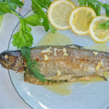 Kwadratowe główne zdjęcie, zbliżenie na soczystego pieczonego pstrąga z kawałkami masła roztopionego na skórce, obok świeży koper i plaster cytryny.