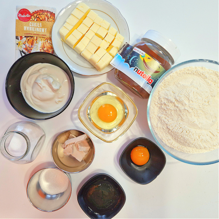 Składniki do przygotowania rogalików krucho drożdżowych ułożone w miseczkach na białym blacie. mąka jajka masło drożdże świeże cukier śmietana 18% cukier waniliowy szczypta soli