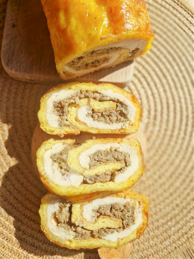 Cała rolada serowa z kurczakiem i pieczarkami ułożona na drewnianej desce. Obok na mniejszej deseczce trzy grube plastry rolady. Tło w odcieniach brązu.