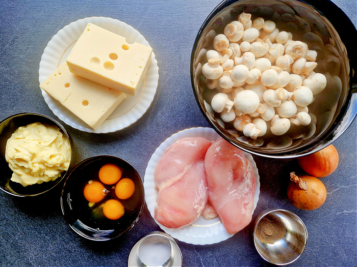 Składniki do przygotowania rolady serowej z pieczarkami na antracytowym blacie ułożone na miskach i talerzach są to pieczarki ser żółty majonez jajka cebula pierś z kurczaka i przyprawy.