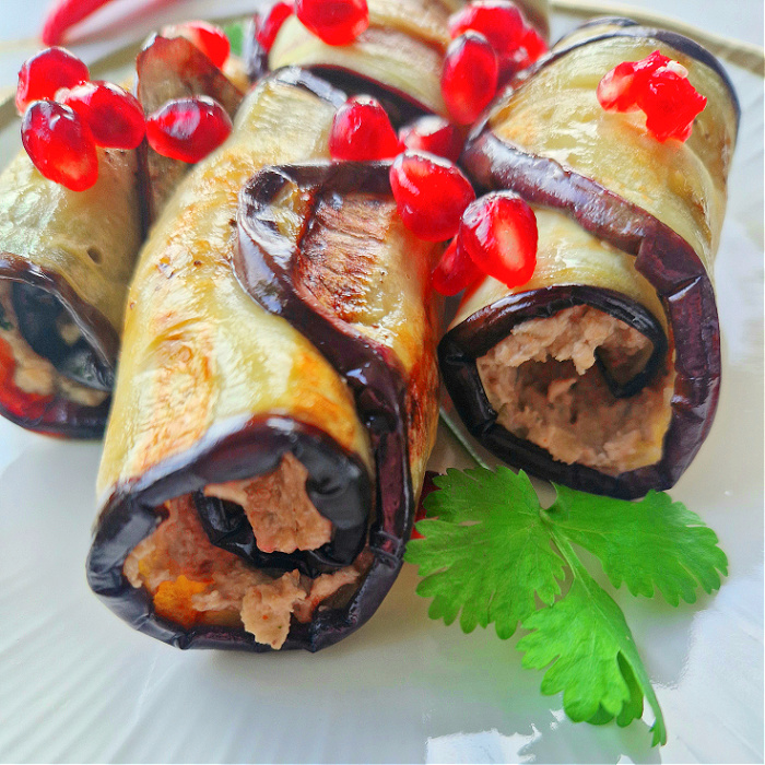 Grillowane roladki bakłażanowe, keto przystawka. Zwinięte z masą orzechowo-serową, ułożone na białym talerzu, przystrojone granatem i zieloną pietruszką.