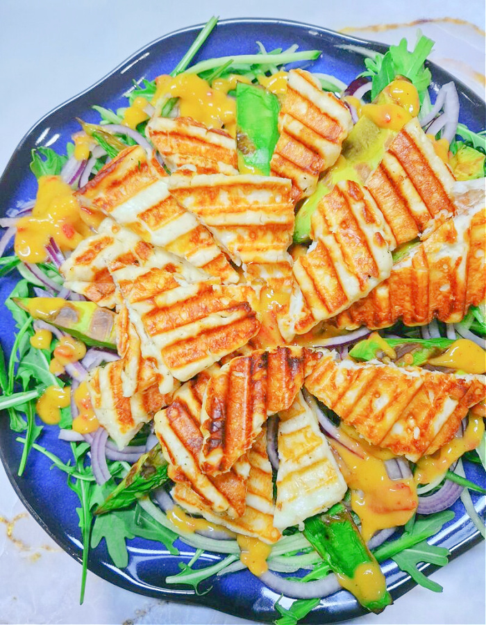 Prostokątne zdjęcie na jasnozłotym tle. Na środku stoi niebieski talerz, a na nim sałatka warstwowa: rukola, czerwona cebula, grillowane awokado, halloumi i polewa z mango-chili.