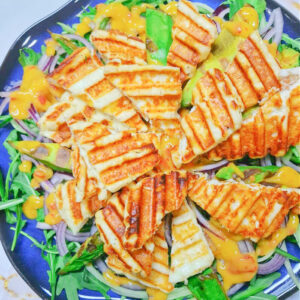Kwadratowe zbliżenie na niebieski talerz z keto sałatką. Widać rukolę, plastry grillowanego sera halloumi, kawałki awokado i pomarańczowy dressing z mango.