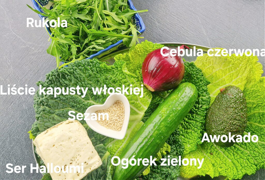 Na antracytowym blacie ułożone składniki sałatki: na półmisku kapusta włoska, obok sezam, ser halloumi, zielony ogórek, awokado, czerwona cebula i rukola.