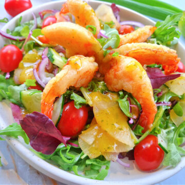 Kwadratowe zdjęcie główne przedstawiające sałatkę z krewetkami w tempurze i mango. Na jasnym talerzu krewetki w złocistej panierce, owoce i świeże zioła na eleganckim biało-złotym tle.