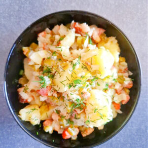 Kwadratowe ujęcie przedstawiające czarną miseczkę z sałatką ziemniaczaną. Widać kawałki ziemniaków, podsmażony boczek, cebulę i ogórki kiszone. Całość posypana świeżym koperkiem. Tło antracytowe, jednolite.