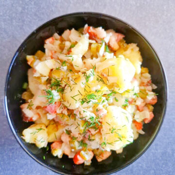 Kwadratowe ujęcie przedstawiające czarną miseczkę z sałatką ziemniaczaną. Widać kawałki ziemniaków, podsmażony boczek, cebulę i ogórki kiszone. Całość posypana świeżym koperkiem. Tło antracytowe, jednolite.