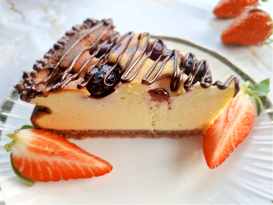 Zbliżenie na kawałek keto sernika z borówkami na białym talerzyku. Obok czerwone truskawki