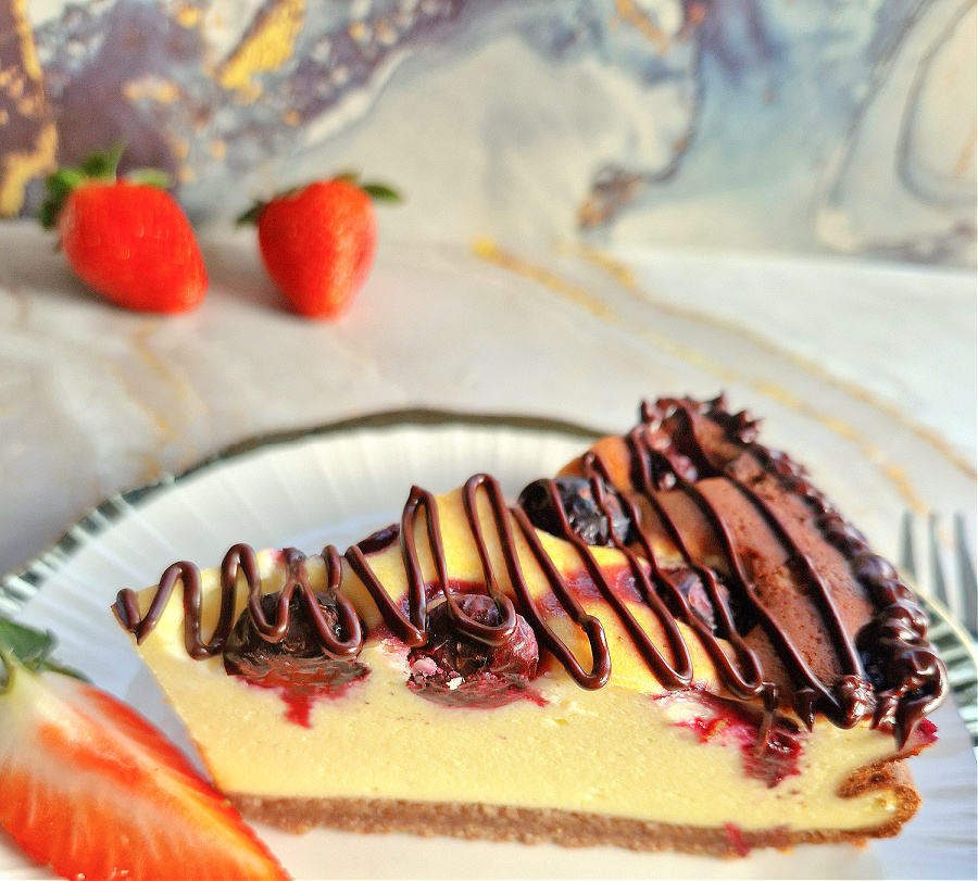 Zbliżenie na trójkątny kawałek keto sernika z borówkami, polanego roztopioną gorzką czekoladą. jasne tło, ciasto na białym talerzyku